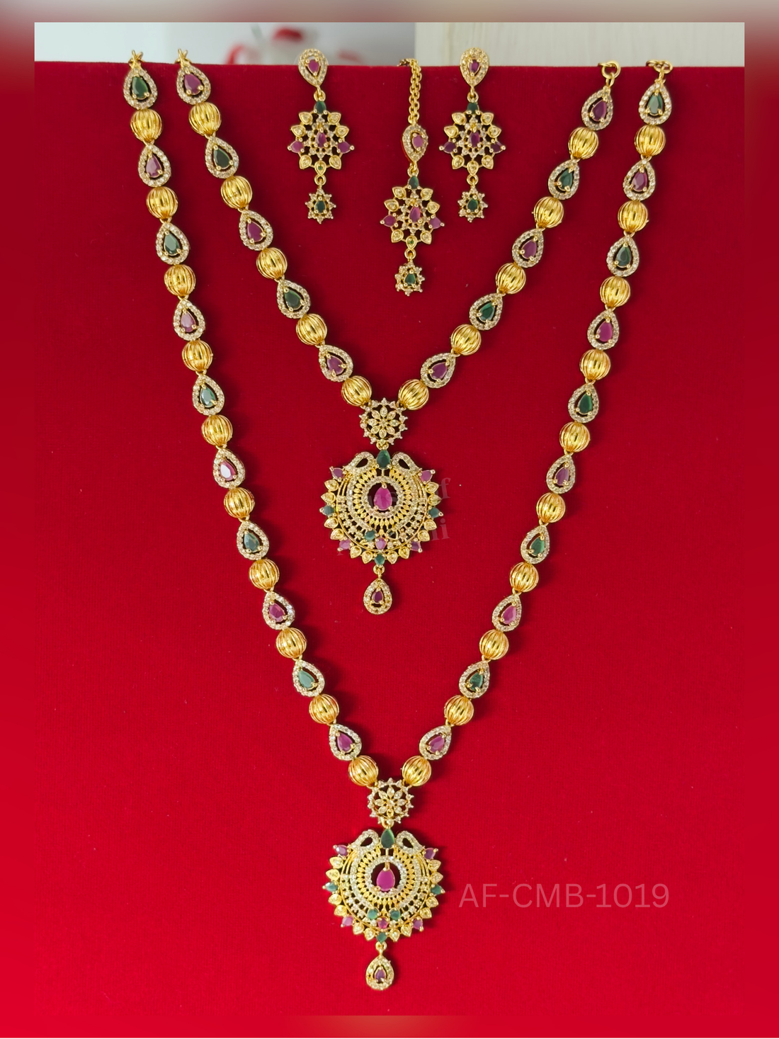 Ribbed gold beads alternate with ruby and emerald teardrops, centred on a round peacock pendant with a dainty drop, and paired with matching earrings and maang tikka.