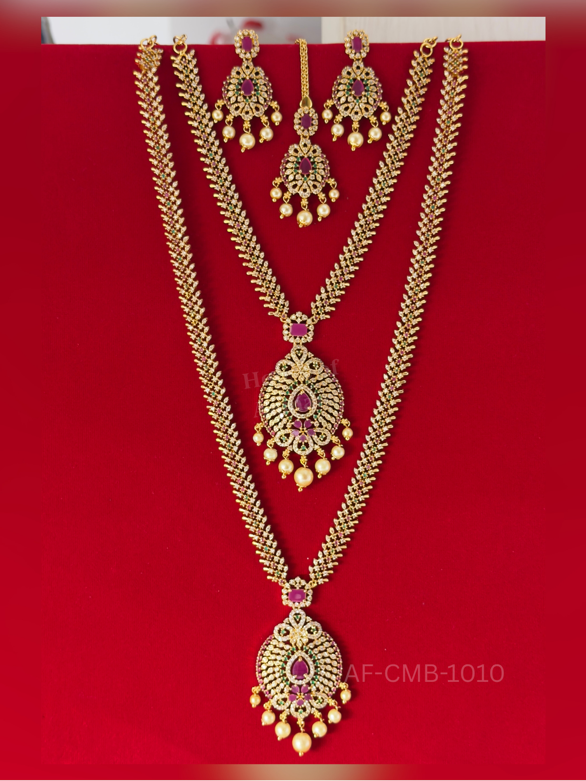 A delicate leaf-patterned chain of ruby, emerald, and white stones ends in a lavish oval pendant with pearl drops, styled with earrings and maang tikka.