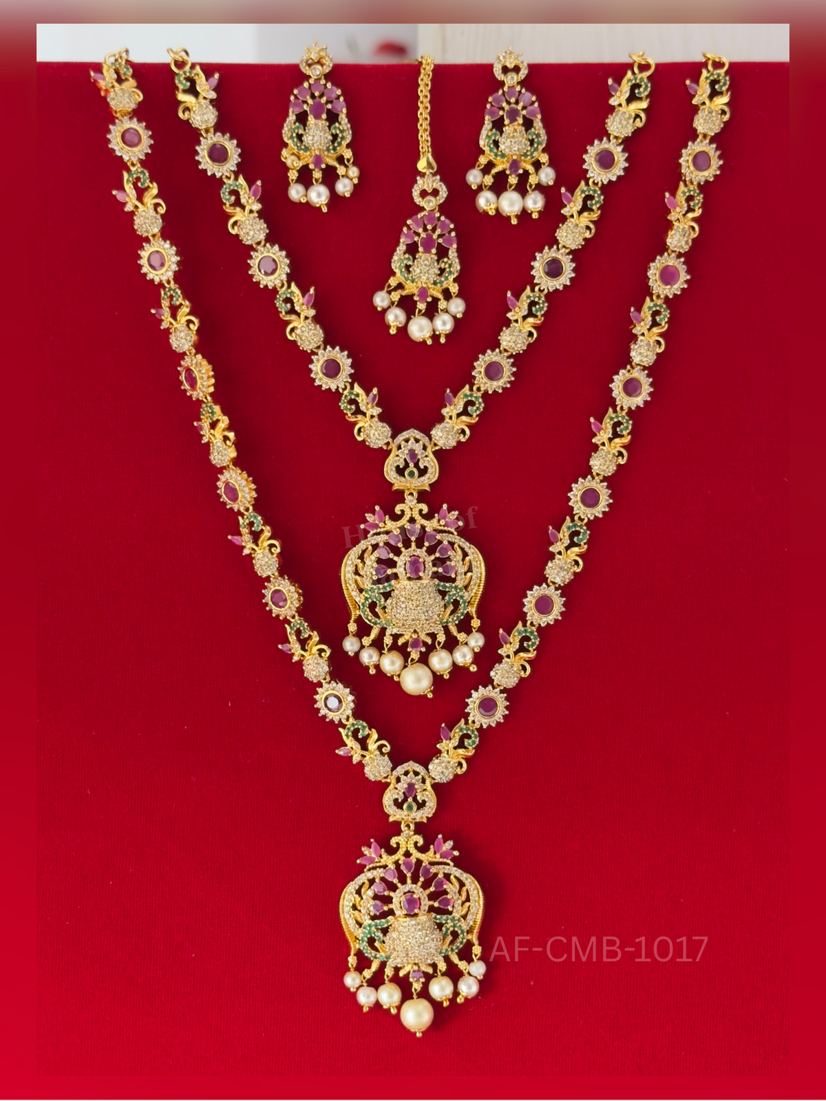 Sunburst ruby stones and floral scroll motifs precede an ornate pendant of ruby petals and emerald curves with pearl drops, plus matching earrings and maang tikka.