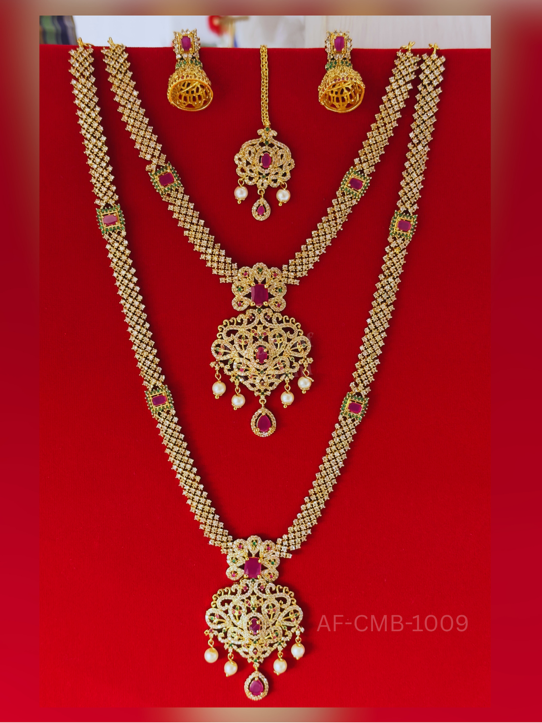 A shimmering lattice chain of white crystals with ruby and emerald accents leads to an intricate scrollwork pendant, paired with jhumka earrings and maang tikka.