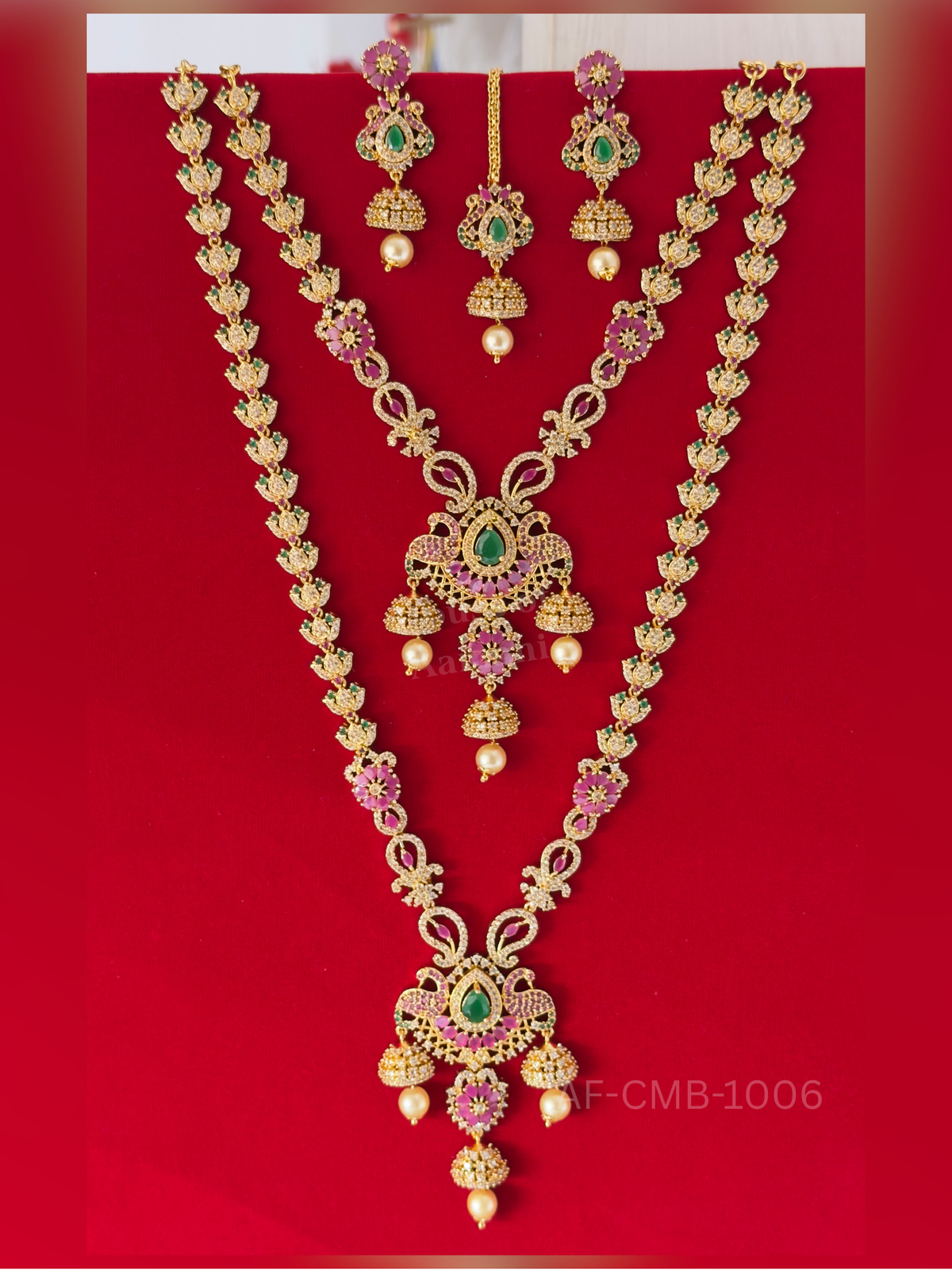 Lotus-shaped links and ruby flower motifs lead to a peacock pendant with an emerald centre and tiered jhumka drops, with matching earrings and maang tikka.