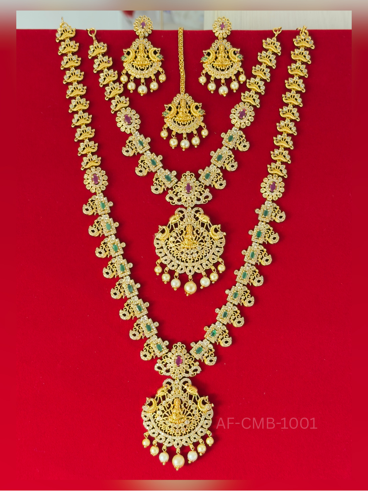 A regal long haram and short necklace featuring Goddess Lakshmi pendants flanked by peacocks, with emerald and ruby accents, pearl drops, and matching earrings and maang tikka.
