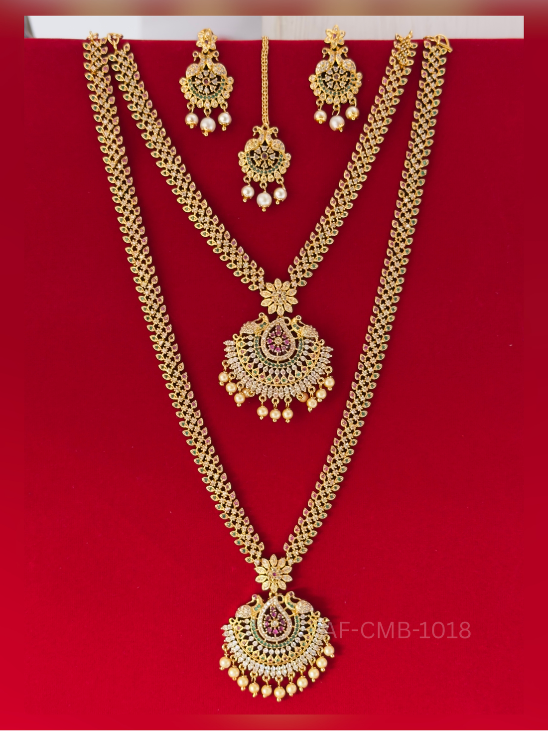 A leaf-patterned chain of white, ruby, and emerald stones supports a peacock chandbali pendant with pearl drops, coordinated with earrings and maang tikka.