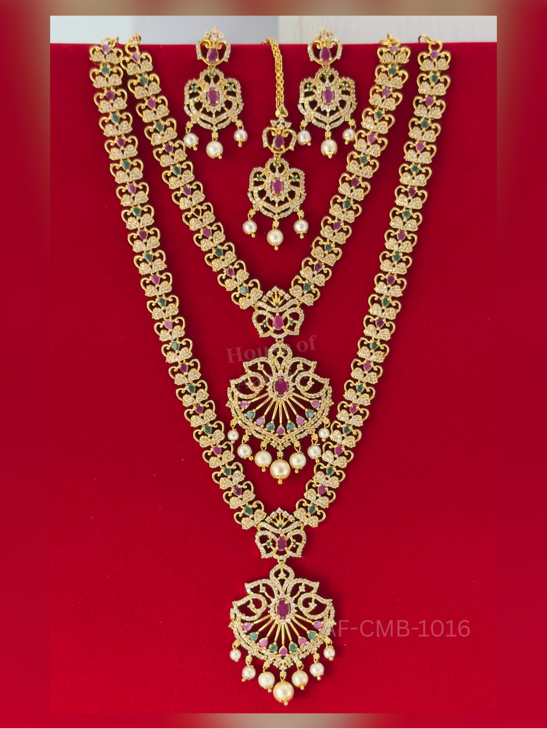 Heart-scroll links in ruby, emerald, and white stones lead to a fan-shaped pendant with pearl drops, styled with earrings and maang tikka.