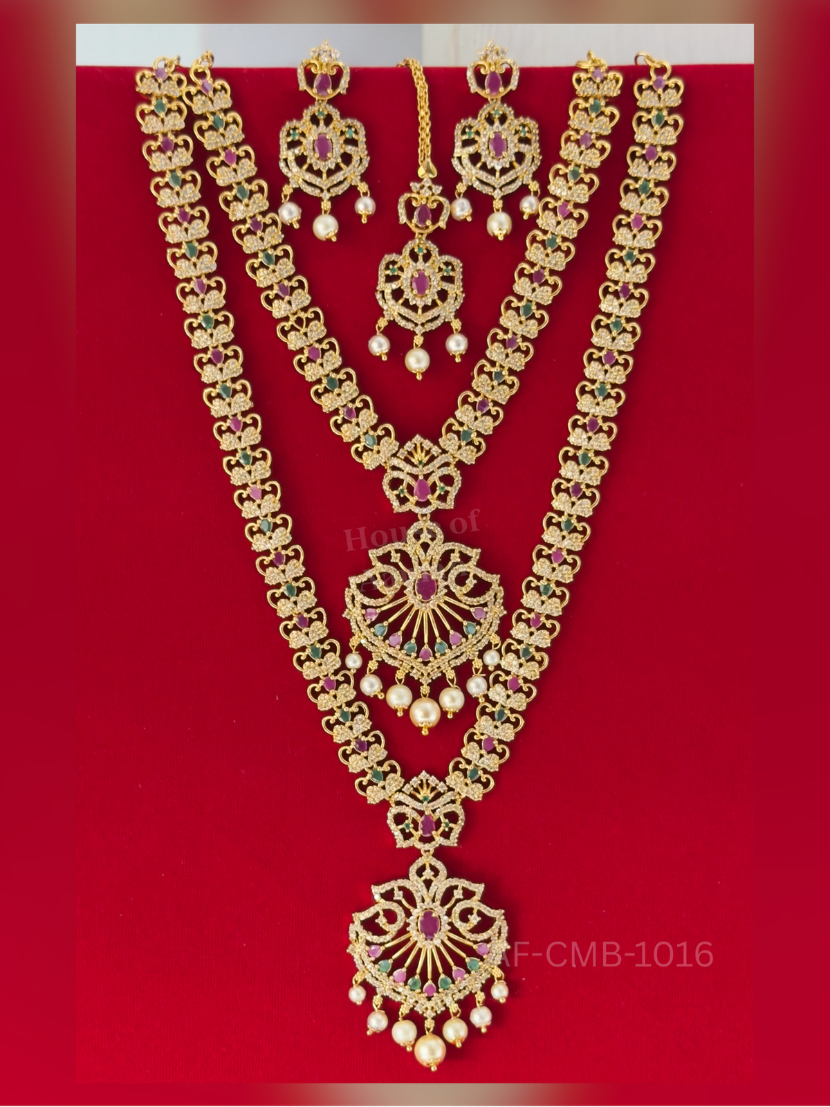 Heart-scroll links in ruby, emerald, and white stones lead to a fan-shaped pendant with pearl drops, styled with earrings and maang tikka.